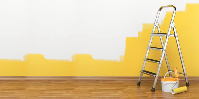 Partially painted wall in yellow with a ladder, paint bucket, and roller tray on a wooden floor by a Boston contractor.