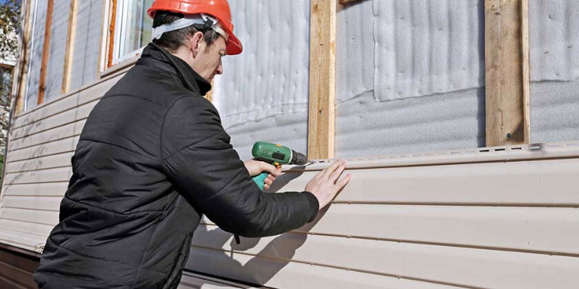 A contractor in a hard hat using a drill on a building's exterior siding.